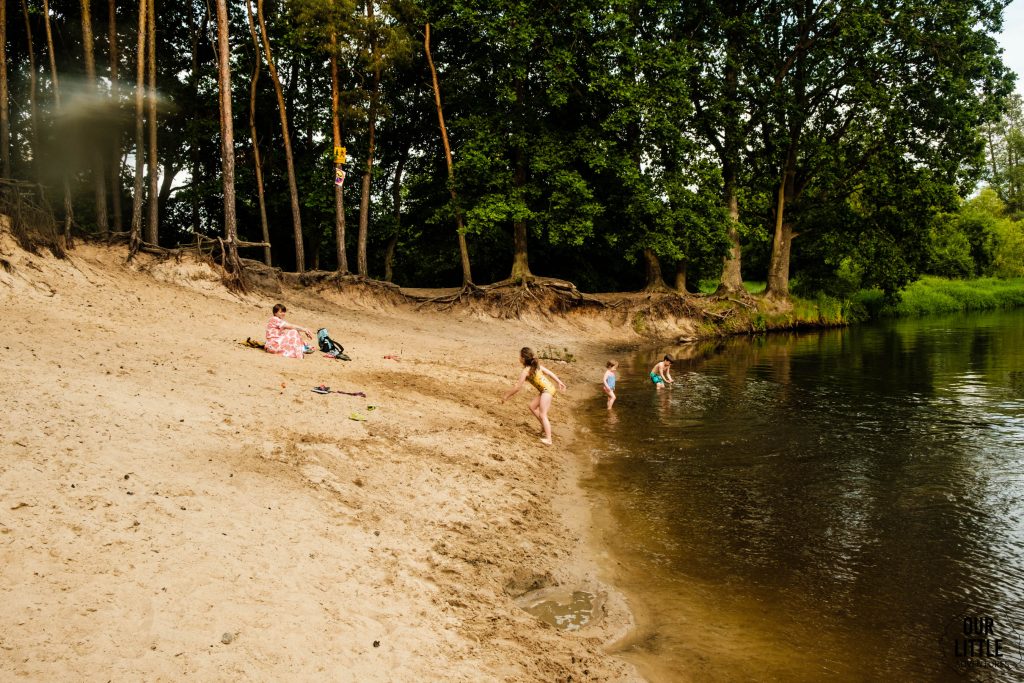 Dzieci kąpią się w leśnej rzece, kobieta siedzi na brzegu. Bezpieczne wakacje z dziećmi, zdjęcie autorstwa OurLittleAdventures.pl