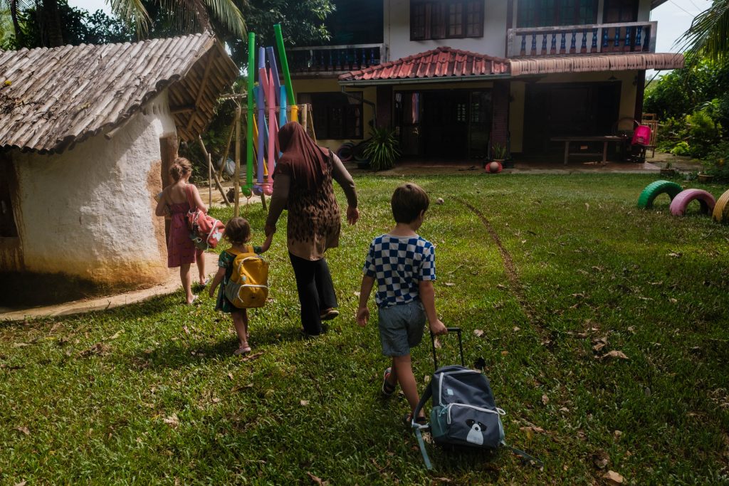 Trójka dzieci idzie po trawie przed tajską szkołą na Koh Lancie, edukacja domowa dzieci, fot. Ourlittleadventures.pl