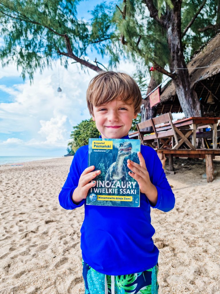 Chłopiec stoi na plaży i trzyma książkę o dinozaurach, zdjęcie pochodzi z tekstu Dinozaury i wielkie ssaki - prawdopodobnie najlepsza książka o dinozaurach dla dzieci ever! na stronie OurLittleAdventures.pl