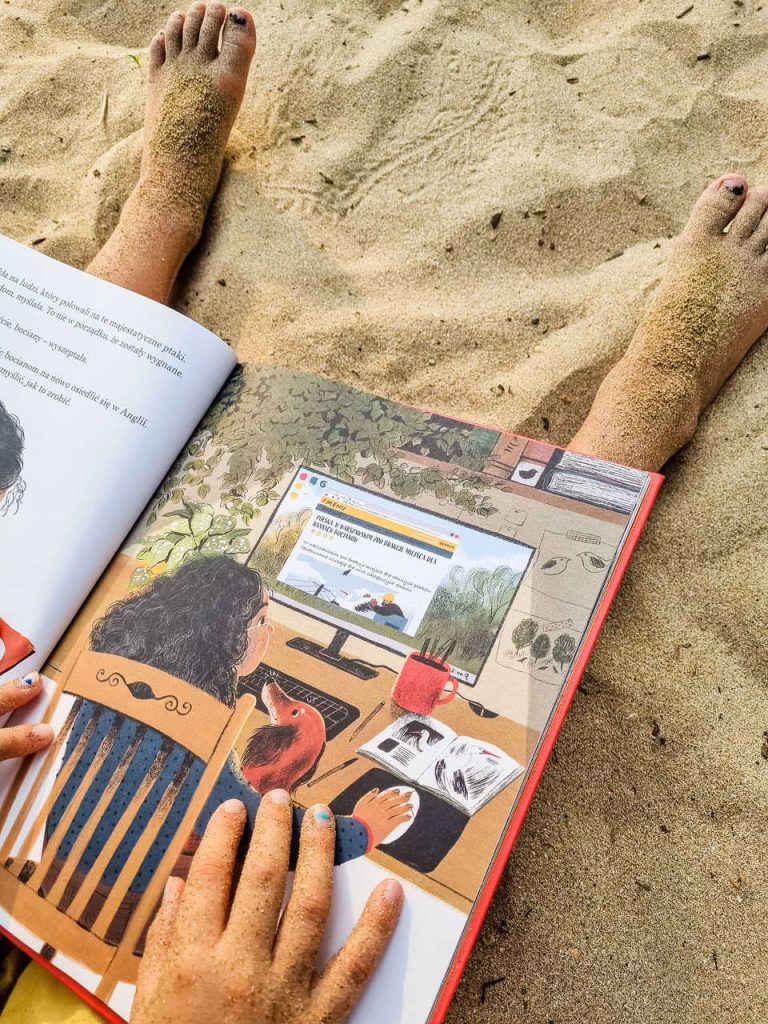 Książka "Kiedy bociany wróciły do domu" wydawnictwo Świetlik trzymana przez chłopca siedzącego na piasku, zdjęcie pochodzi ze strony OurLittleAdventures.pl - książki przyrodnicze dla dzieci