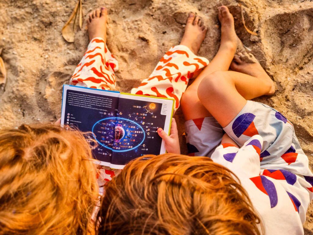 Dzieci czytają na plaży książkę "Jak naukowcy mierzą świat", Darek Aksamit, zdjęcie pochodzi z tekstu na blogu ourlittleadventures.pl - Pytaj, sprawdzaj, eksperymentuj razem z dzieckiem, nawet jeśli nie znasz odpowiedzi - rozmowa z Darkiem Aksamitem, naukowcem i popularyzatorem nauki