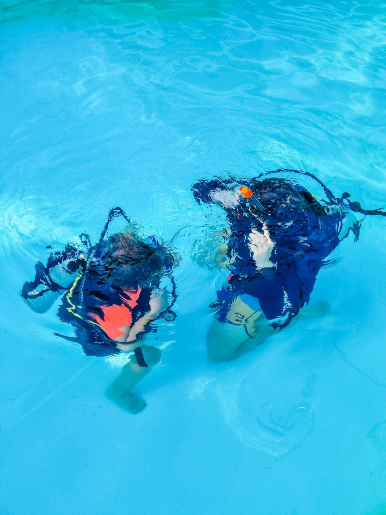 Dwie osoby zanurzają się z butlą w basenie - zdjęcie pochodzi z bloga OurLittleAdventures.pl z tekstu: Jak zacząć nurkować? - wszystko, co musisz wiedzieć o nurkowaniu