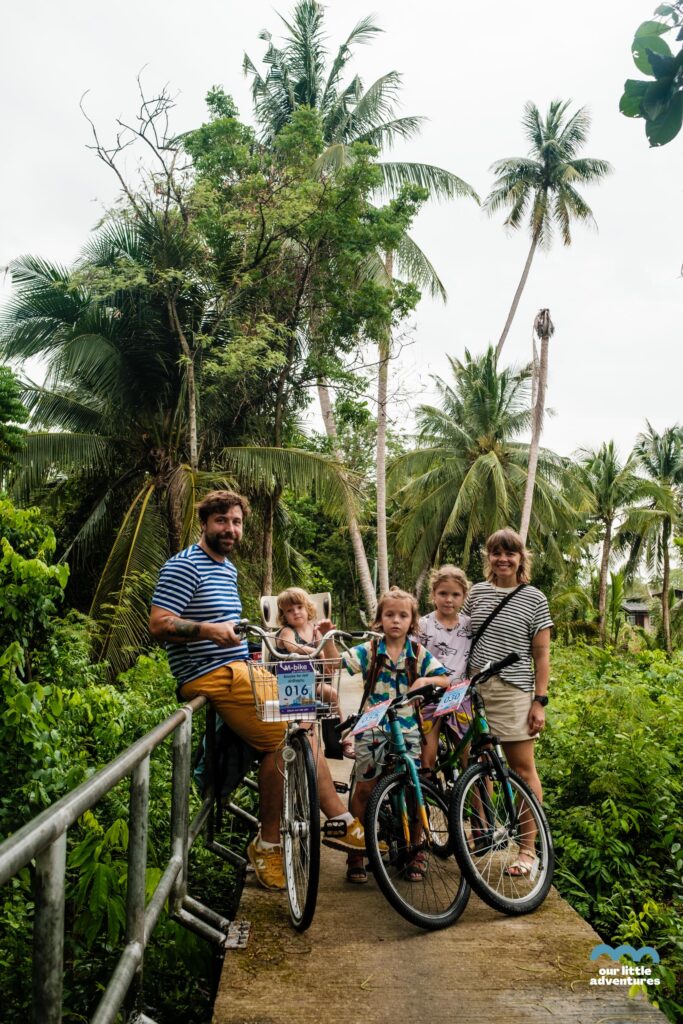 Rodzina z dziećmi na rowerach na wąskiej kładce rowerowej w Azji - zdjęcie pochodzi z tekstu na blogu OurLittleAdventures.pl pt. Wyspa Bang Krachao - wycieczka rowerowa z dziećmi do zielonej oazy Bangkoku